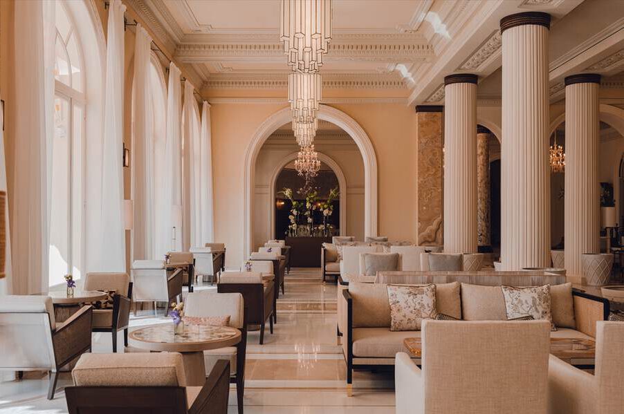 Geräumige, elegante Lounge mit cremefarbenen Sofas, runden Holztischen, hohen weißen Säulen, großen Bogenfenstern und Kronleuchtern, die von der Decke hängen.