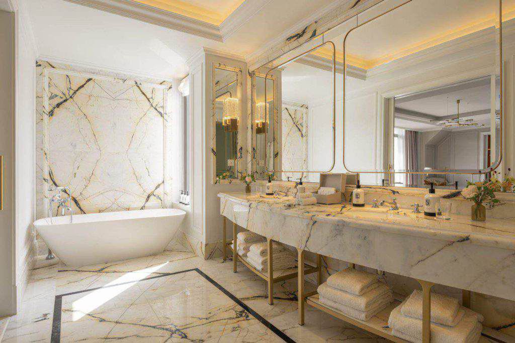 Luxuriöses Badezimmer mit Marmorböden und -wänden, einem großen Waschtisch mit zwei Waschbecken, einer freistehenden Badewanne, Spiegeln und ordentlich angeordneten Handtüchern und Toilettenartikeln.