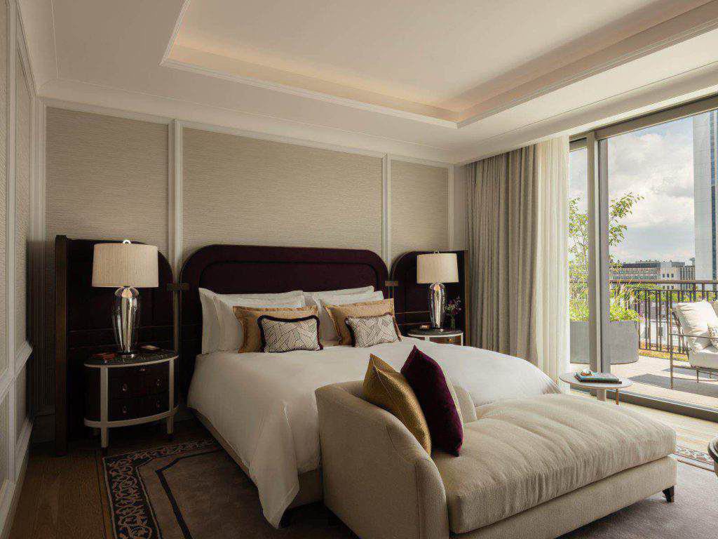 Ein modernes Schlafzimmer mit einem großen Bett, zwei Nachttischen mit Lampen, einer gepolsterten Bank am Fußende des Bettes und Glasschiebetüren, die auf einen Balkon führen.