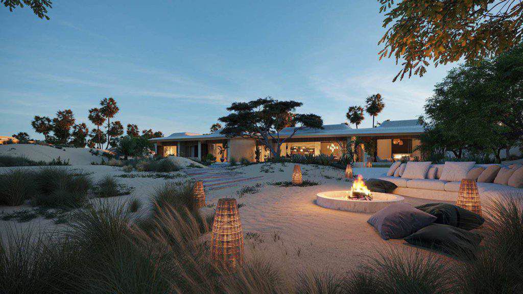 Sitzbereich im Freien mit einer Feuerstelle, umgeben von Sand, Laternen und Kissen, mit einem modernen Haus und Palmen im Hintergrund in der Abenddämmerung.