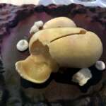 Dessert, das an ein aufgeschlagenes Ei erinnert, mit einer cremigen Füllung auf einem strukturierten schwarzen Teller, elegant garniert mit kleinen Baiserstückchen.