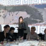 Eine Gruppe von Menschen sitzt an einem Tisch. Auf dem Bildschirm wird eine Luftaufnahme einer Küstenregion projiziert. Eine Frau mit einem Blatt Papier steht neben dem Bildschirm, während TastEmotion mit seiner einzigartigen Mischung aus Innovation und kulinarischer Leidenschaft für die Atmosphäre sorgt.