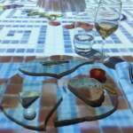 Ein gedeckter Tisch mit einem zerbrochenen Keramikteller, Brot, Käse, einer Kirschtomate und Gläsern mit Weißwein und Wasser auf einer karierten Tischdecke.