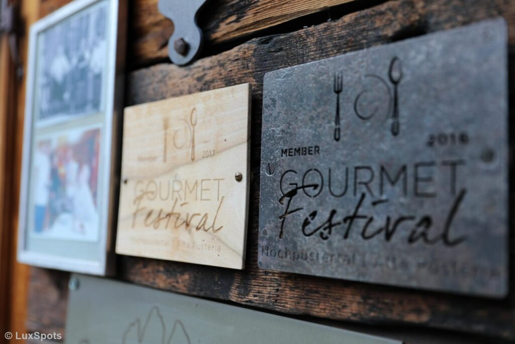 Nahaufnahme von zwei Schildern an einer Holzwand. Auf beiden steht „Gourmet Festival“ und über dem Text sind einfache Bestecksymbole zu sehen. Die Schilder sind aus unterschiedlichen Materialien und in unterschiedlichen Farben.