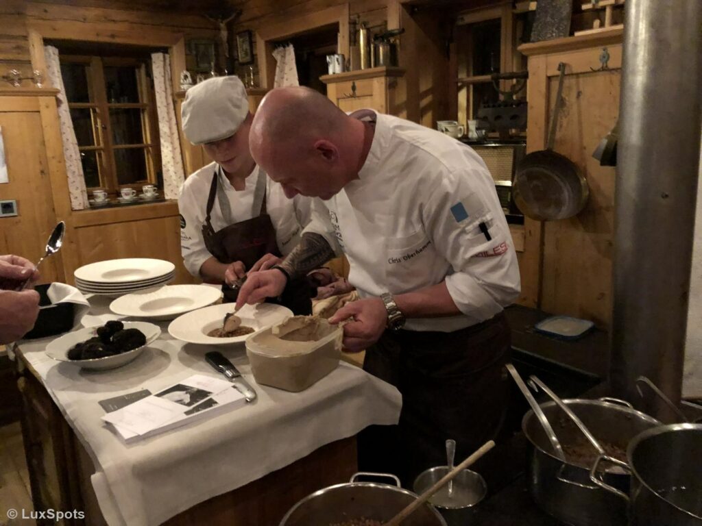 Köche in einer rustikalen Küche richten Gerichte sorgfältig auf einer Holzarbeitsplatte an. Im Hintergrund sind verschiedene Küchenutensilien und Zutaten zu sehen.