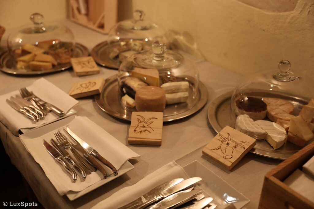 Eine Ausstellung verschiedener Käsesorten auf Tabletts mit Glaskuppeln, begleitet von Holztafeln mit Abbildungen. Silberbesteck ist ordentlich daneben angeordnet.