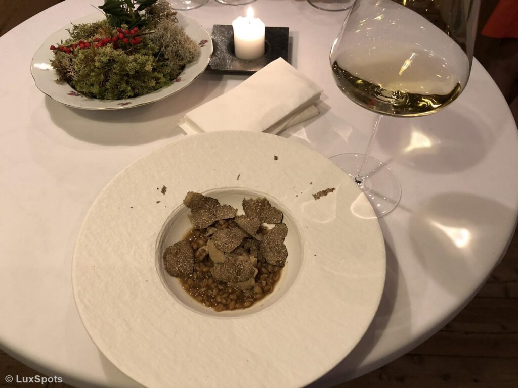 Ein weißer Teller mit Risotto, garniert mit gehobelten Trüffeln, neben einem Glas Weißwein und einer brennenden Kerze auf einem runden Tisch.