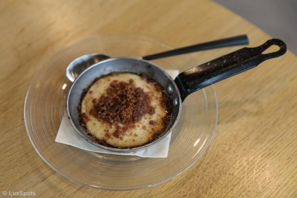 Eine kleine Bratpfanne mit einem Dessert, garniert mit zerbröseltem braunem Topping, auf einem durchsichtigen Glasteller mit einem Löffel und einer Serviette darunter.