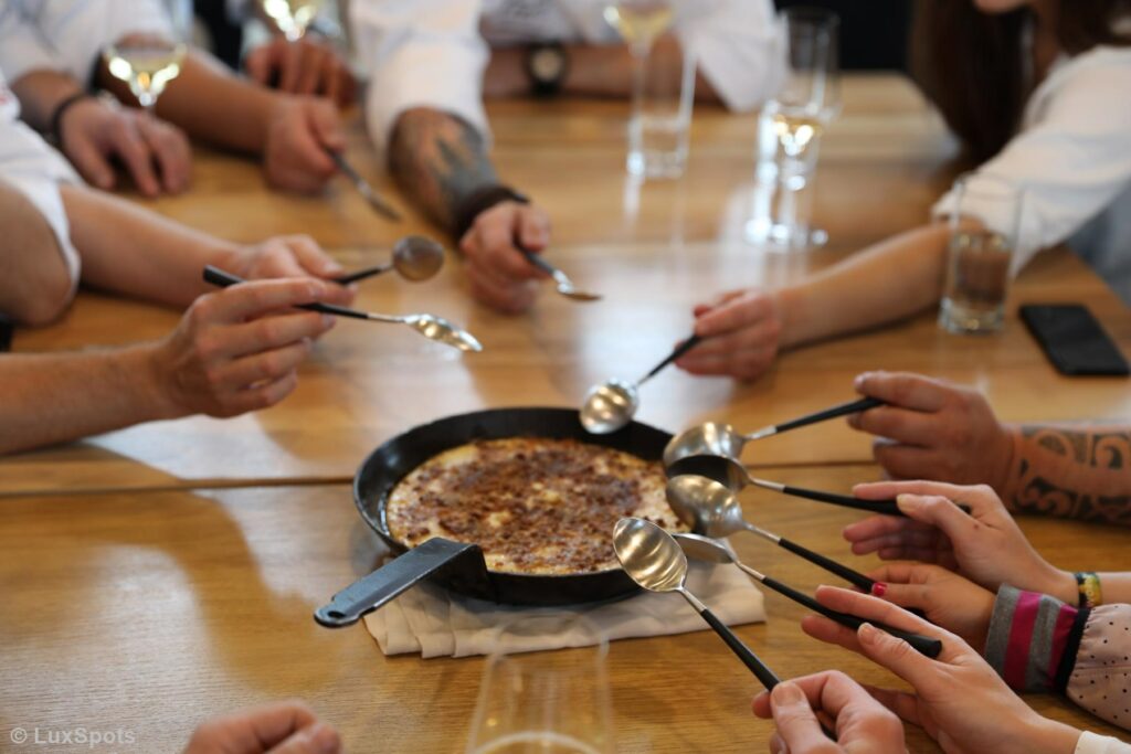 Eine Gruppe von Menschen versammelt sich um einen Tisch. Jeder hält einen Löffel in der Hand und greift nach einem Dessert in einer Bratpfanne.