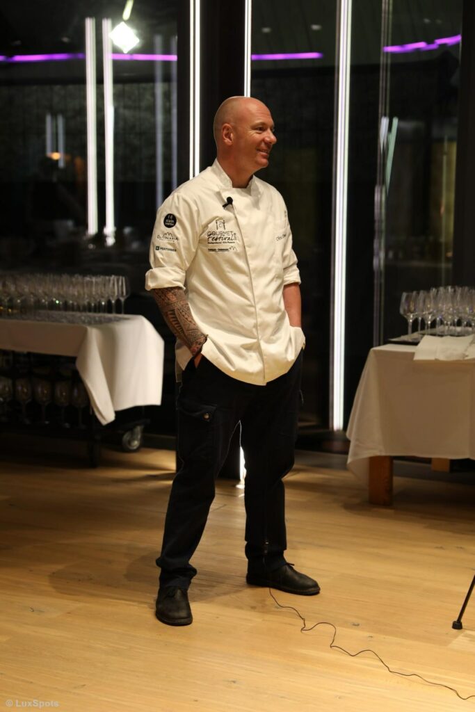 Ein Koch in weißer Uniform steht in einem Restaurant oder Veranstaltungsort und beantwortet Fragen für ein Videointerview.