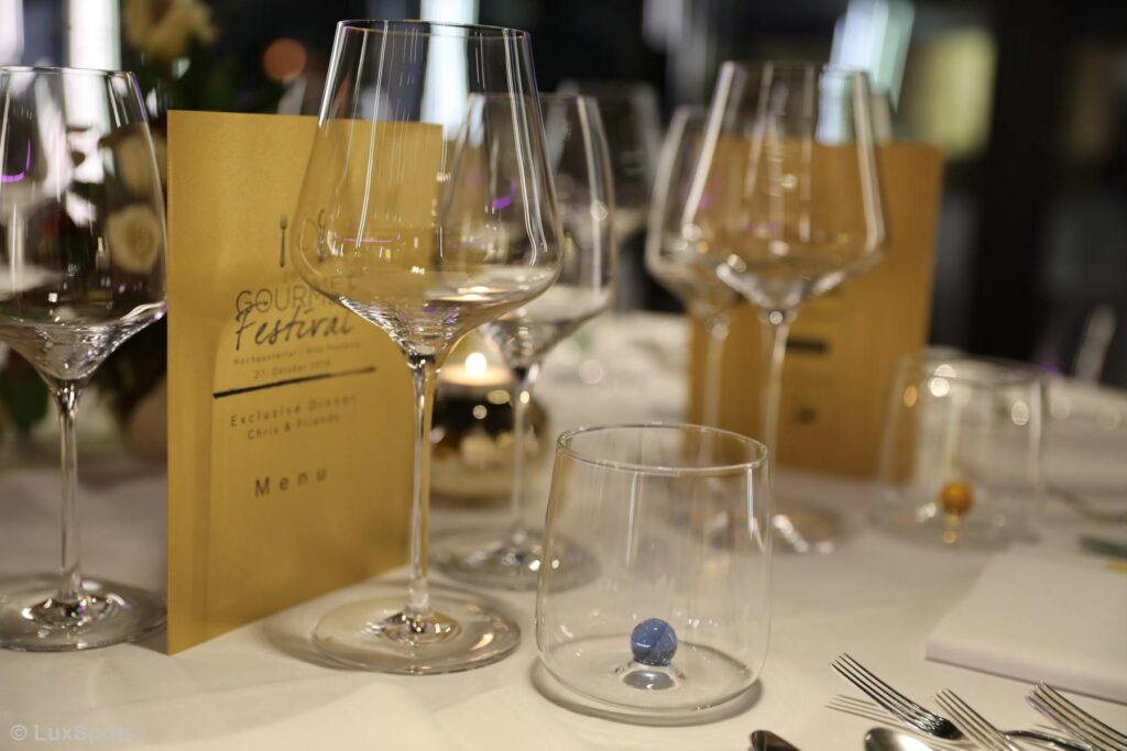 Elegant gedeckter Tisch mit Weingläsern, einer Kerze und einer Speisekarte auf einer weißen Tischdecke, vorbereitet für ein feines kulinarisches Erlebnis.