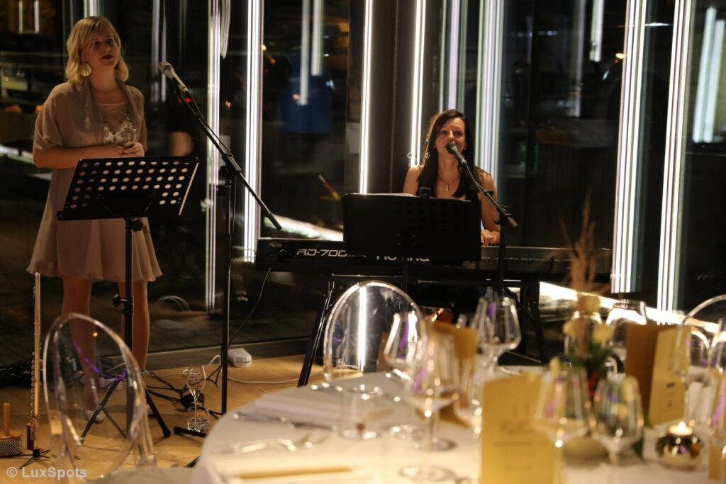 Zwei Musikerinnen spielen im Hintergrund bei einem Abendessen; eine spielt ein Keyboard, die andere singt mit einem Mikrofon. Im Vordergrund steht ein gedeckter Esstisch.