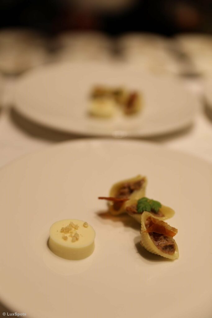 Ein weißer Teller mit Gourmet-Muscheln aus Nudeln, einer kleinen Garnitur und einem runden Stück Käse mit Salzflocken.