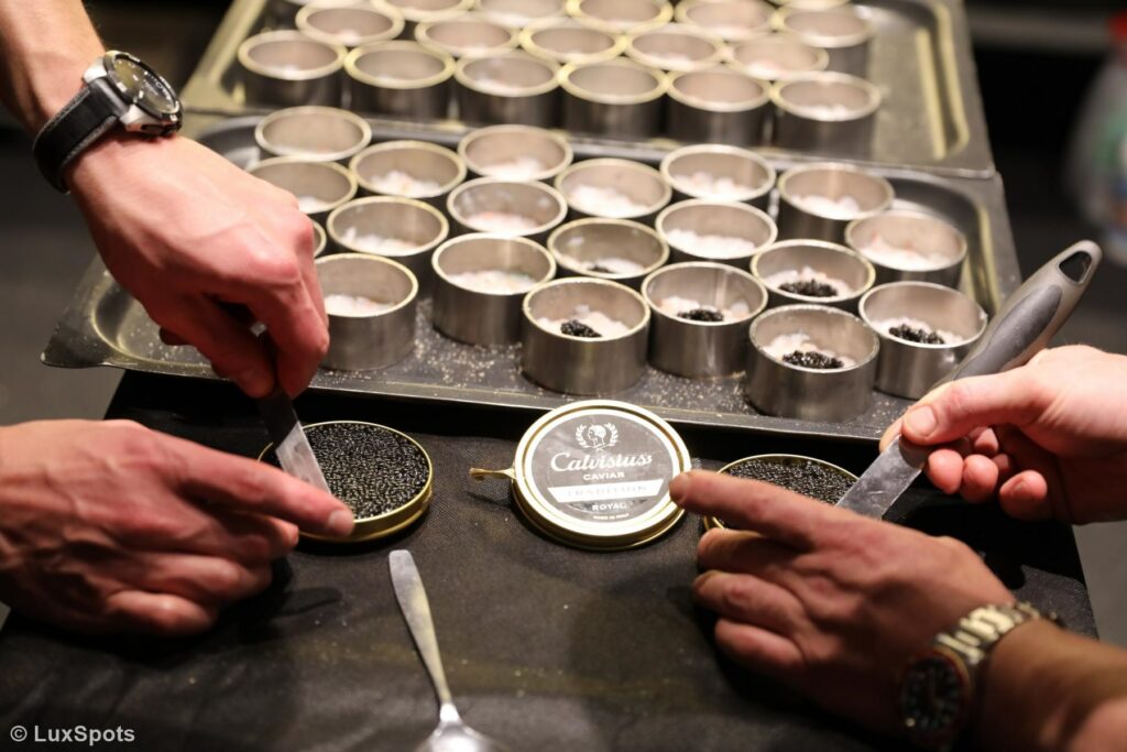 Zwei Personen richten mit Messern Kaviar in mehreren Anrichteformen auf einem Tablett zu. In der Mitte steht ein Deckel mit der Aufschrift „Calvisius-Kaviar“.