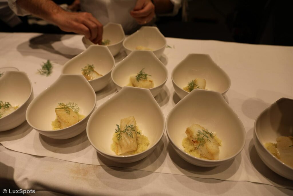 Mehrere weiße Schüsseln auf einem Tisch, jede mit einem Stück Fisch auf einem Bett aus Fenchelpüree, garniert mit Dill. Eine Person garniert eine Schüssel.