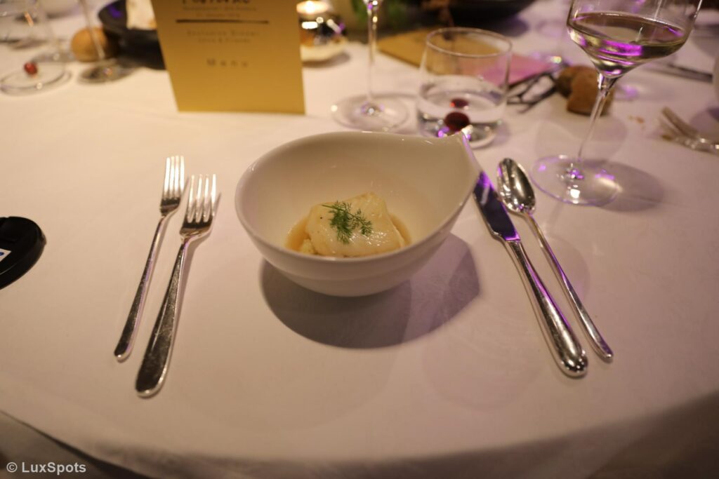 Eine Schüssel mit garniertem Essen steht auf einer weißen Tischdecke, umgeben von Besteck, Gläsern und einer Menükarte.