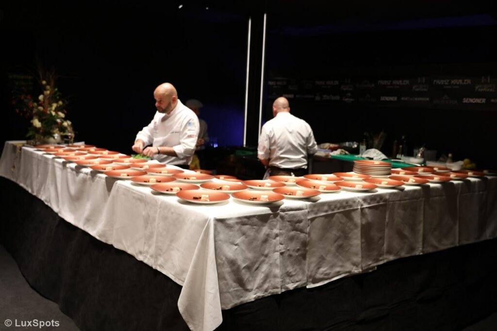 Zwei Köche bereiten in einem schwach beleuchteten Raum auf einem langen, mit einer weißen Tischdecke bedeckten Tisch zahlreiche identische Tellergerichte zu.