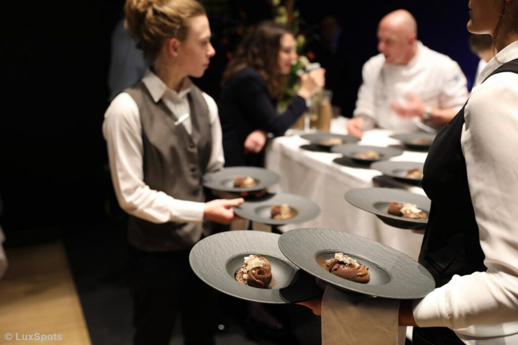 Kellner in Uniform halten Teller mit Desserts, während im Hintergrund Leute an einem Tisch sitzen.