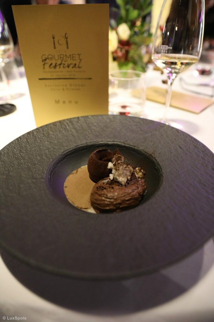 Ein Gourmet-Dessert auf einem stilvollen schwarzen Teller, gedeckt auf einem Tisch mit einer Speisekarte und einem Weinglas im Hintergrund.