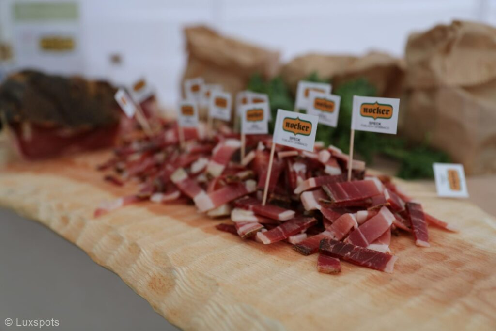 Auf einer Holzplatte angerichtete Scheiben von gepökeltem Fleisch, in einige Stücke sind kleine Fähnchen mit der Aufschrift „Nocker“ eingelegt.