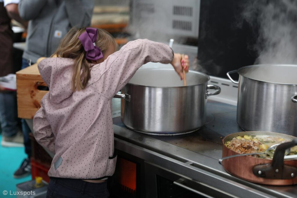 Ein Kind in einer gepunkteten Jacke rührt in einem großen Topf auf dem Herd, Dampf steigt auf. Daneben steht ein weiterer Topf mit Essen.