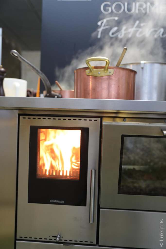 Ein Herd mit sichtbarer Flamme und einem Kupfertopf darauf, vor einem Hintergrund aus Küchengeräten.