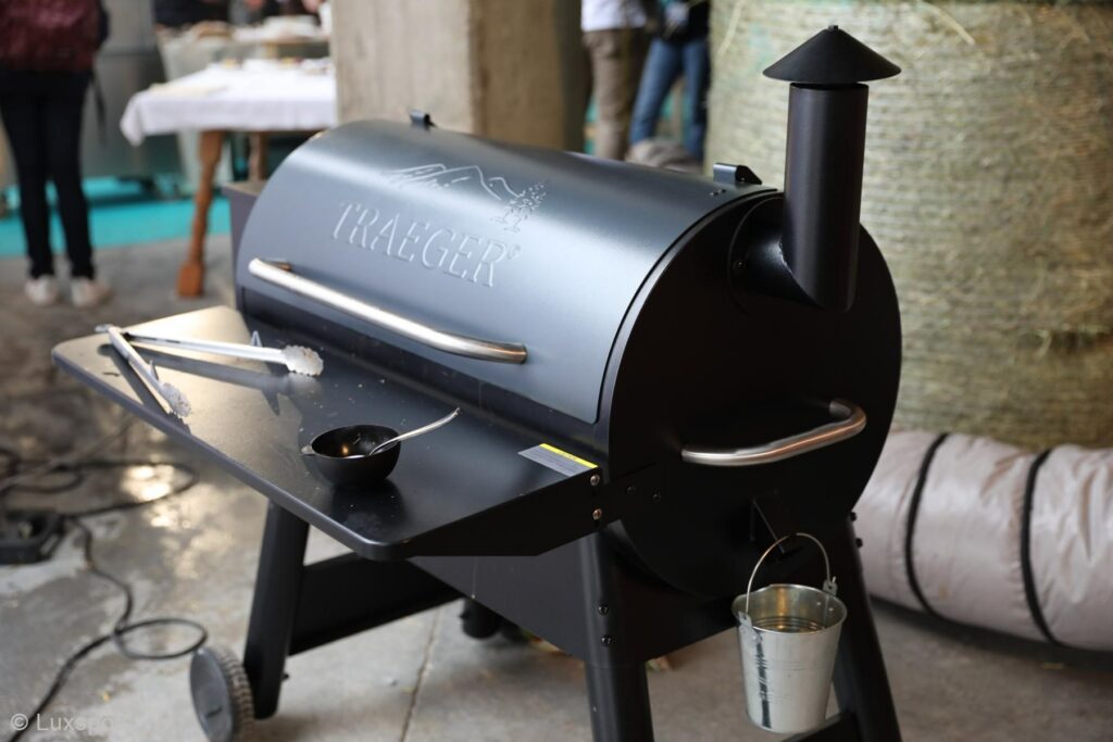 Ausgestellt ist ein Traeger-Grill mit einem kleinen Topf und einer Zange auf der Seitenablage sowie einem Ascheeimer, der an einem Haken hängt.