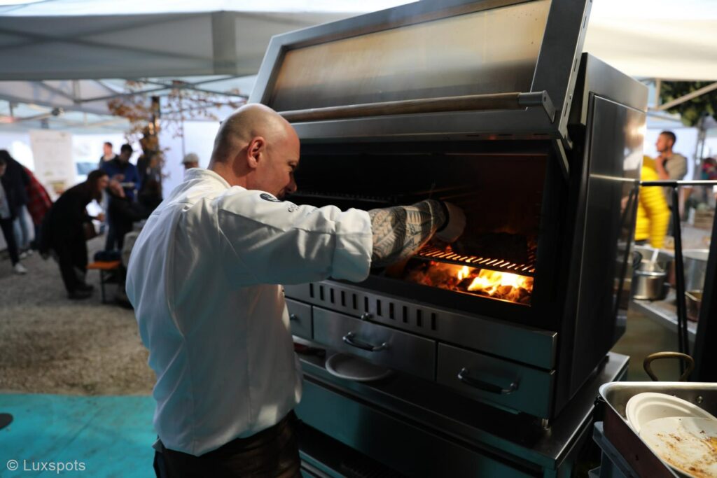 Ein Koch in einer weißen Jacke grillt Essen auf einem Außengrill unter einem Zelt. Im Hintergrund sind andere Personen und Tische zu sehen.