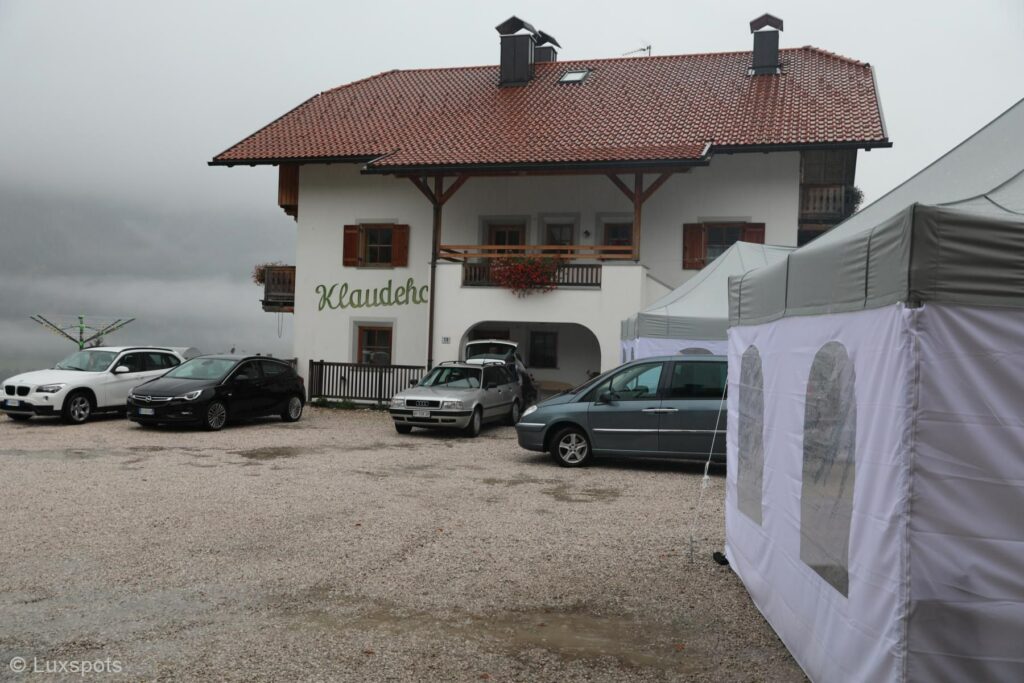 Vor einem Gebäude mit rotem Dach und Balkonen parkende Autos schaffen eine einladende Szene im Hochpustertal. Auf der rechten Seite ist ein Zelt aufgebaut und darüber ragt ein wolkiger Himmel hervor, der den ruhigen Charme der Gegend noch verstärkt.