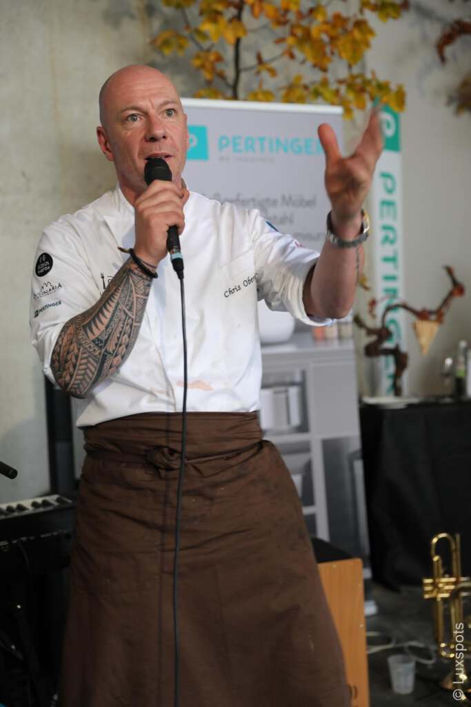 Koch hält eine Rede. Er trägt eine weiße Kochjacke und eine braune Schürze und hält ein Mikrofon. Im Hintergrund sind Herbstblätter und ein Schild mit der Aufschrift „PERTINGER“ zu sehen.