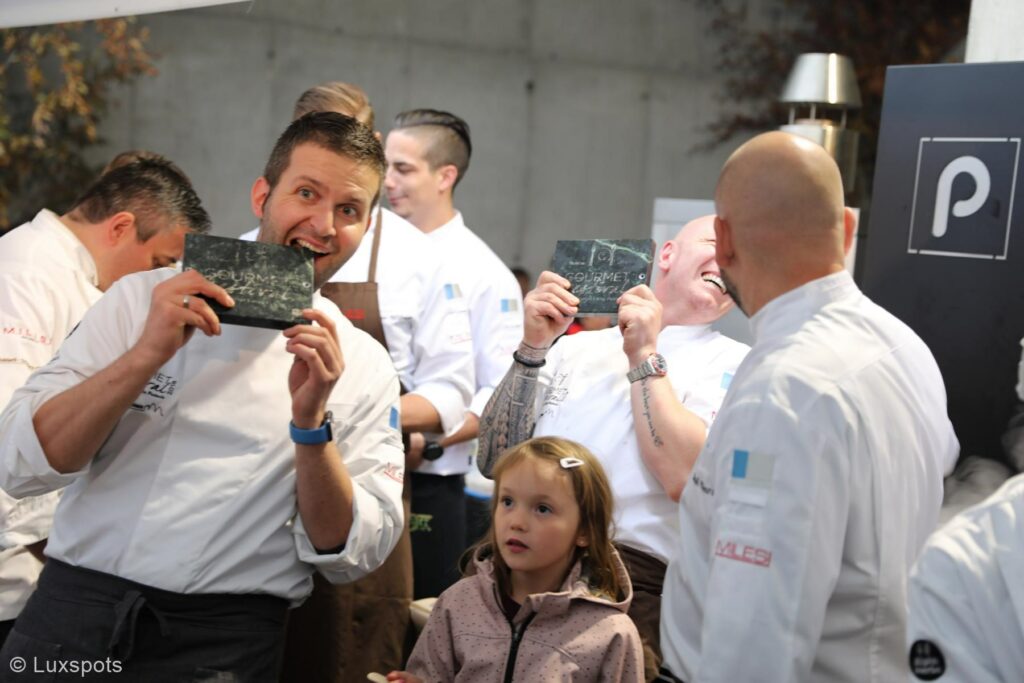 Köche in weißen Uniformen halten spielerisch Schiefertafeln mit der Gravur "Gourmet Festival 2018" hoch, während ein Kind davor steht und zusieht.