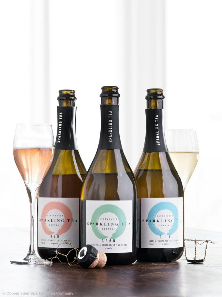 Drei Sektflaschen stehen geöffnet nebeneinander auf einem Holztisch. Links ist ein rosefarbenes Sektglas und rechts im Hintergrund ein gelbliches Sektglas zu sehen. Vor den Flaschen liegen Sektkorken. Auf den Etiketten der Flaschen steht jeweils "Sparkling Tea" geschrieben.
