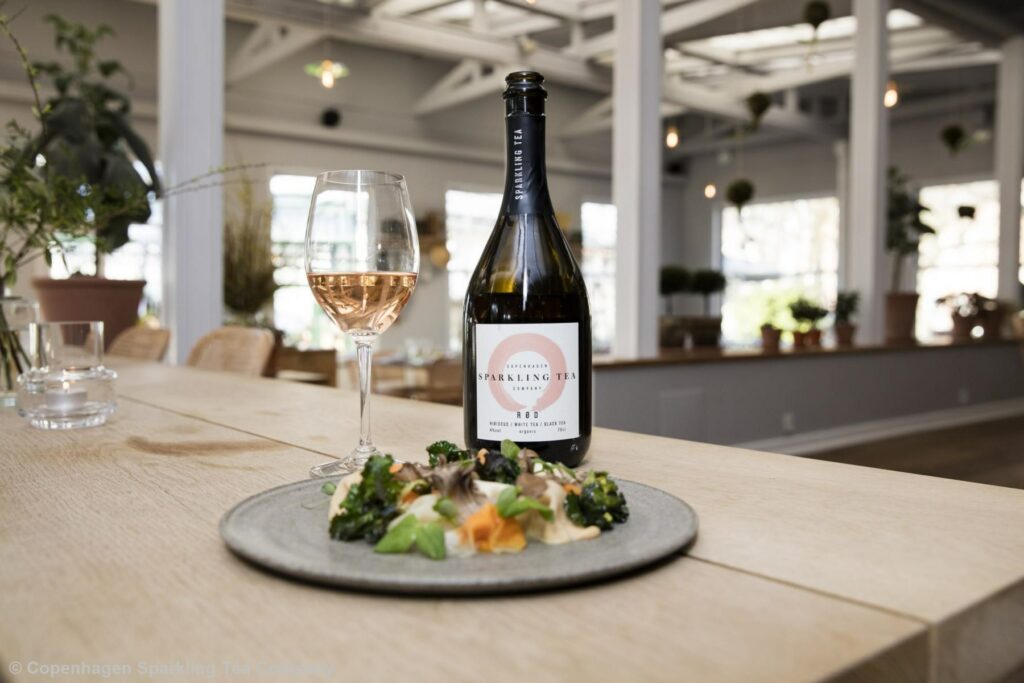 Eine Sektflasche steht mit einem rosefarbenen, alkoholfreien Sekt auf einem Tisch. Auf der Flasche steht "Sparkling Tea".
