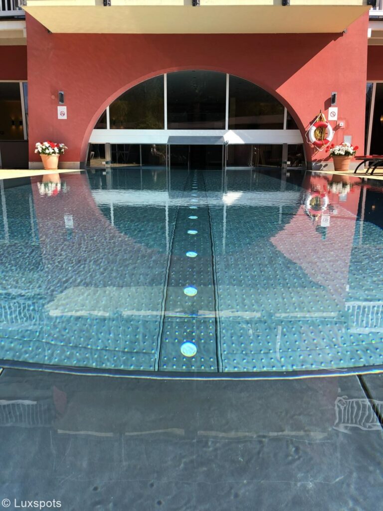 Ein Außenpool mit Blick auf einen halbrunden, gläsernen Eingang zum Innern eines Innenpools eines Hotels in Seefeld, Tirol.