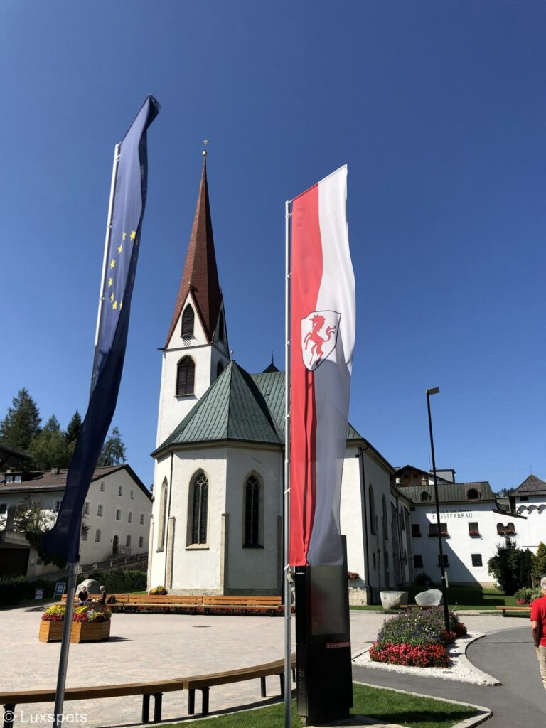 Dorfplatz mit Blumen und einer weiß angestrichenen Kirche und rotem, spitzen Dach des Kirchturms in Seefeld, Tirol. Davor sind die beiden Flaggen Tirols und Europas zu sehen.