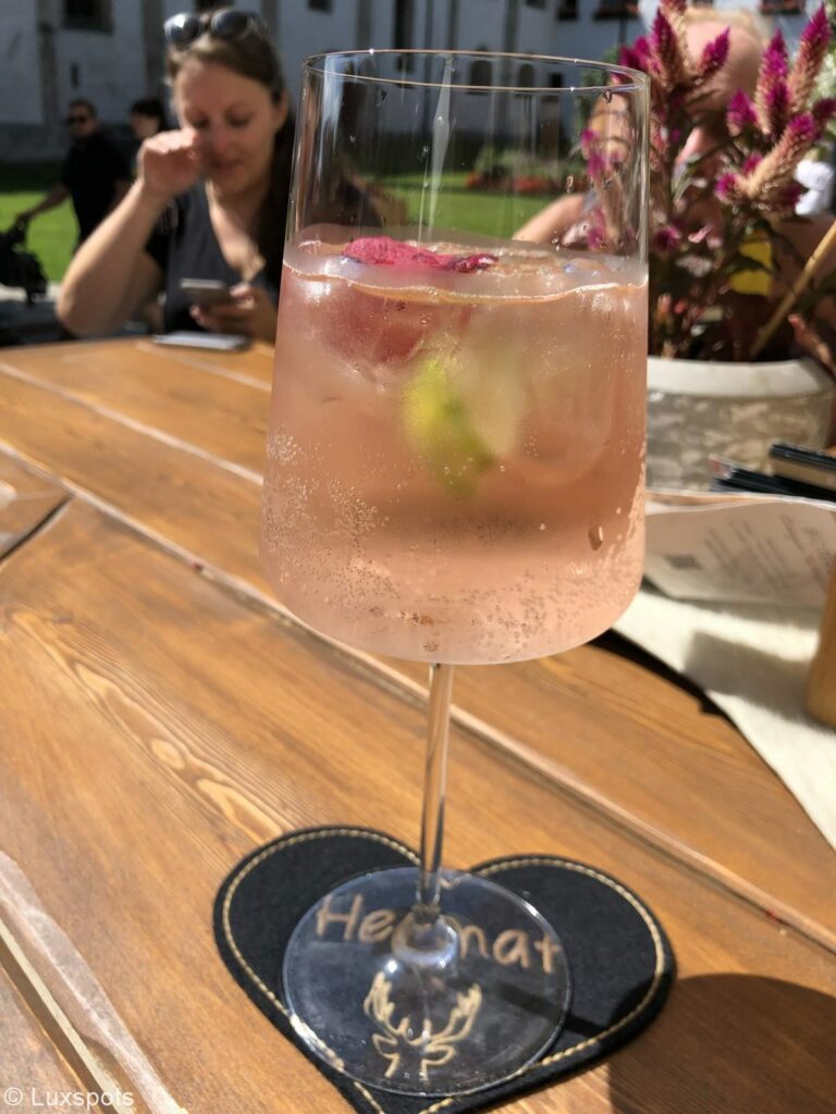 Ein roséfarbener Cocktail mit Heimatbierdeckel steht auf einem Holztisch im Freien. Darin schwimmen Eiswürfel und Limetten.
