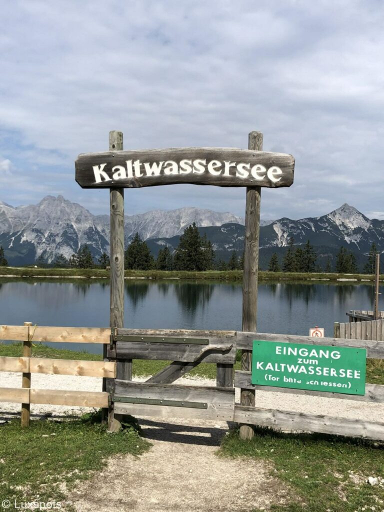 Schild mit der Aufschrift "Kaltwassersee" in Seefeld, Tirol. Dahinter sind der See und viele Berge zu sehen.