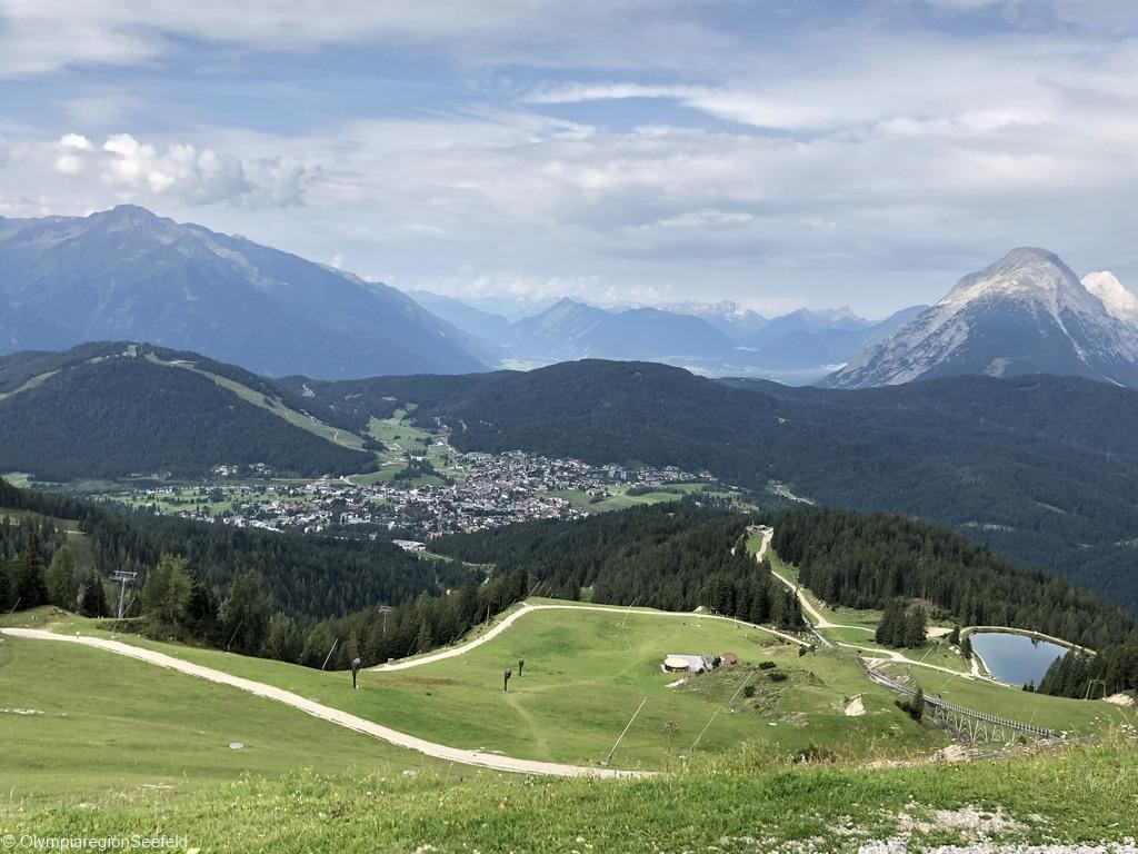 Blick auf das sommerliche Hochplateau Seefeld, mit grünen Skipisten und einem kleinen See für die Beschneiungsanlagen im Winter. Im Hintergrund sind weitere Gebirge der Alpen zu sehen.