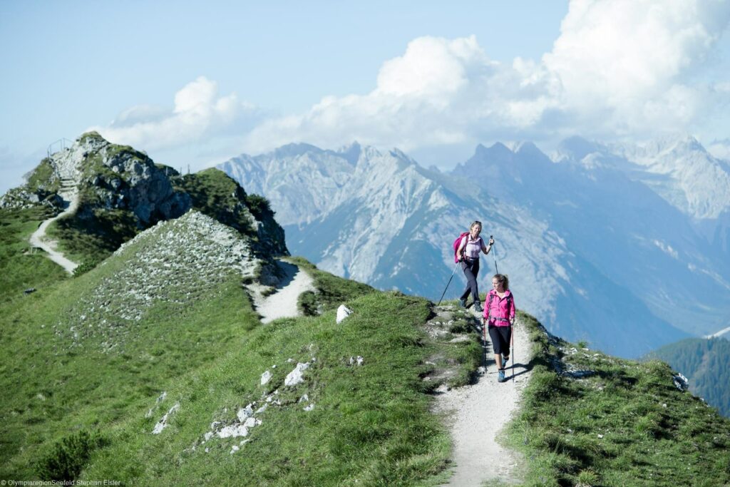 Zwei Frauen wandern mit Wanderstöcken und Rucksäcken auf dem Karwendel Gebirge in Tirol. Im Hintergrund sind weitere Berge udn Wolkenformationen zu sehen.