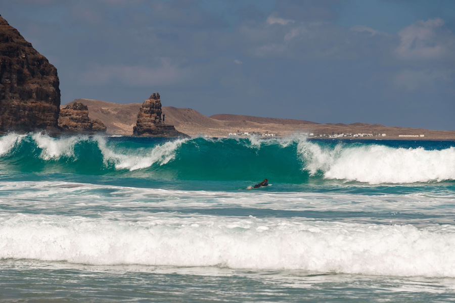 Ein Surfer reitet an einem sonnigen Tag auf einer türkisfarbenen Welle in der Nähe der Felsklippen von Playa de la Canteria – perfekt zum Wellenreiten auf Lanzarote.