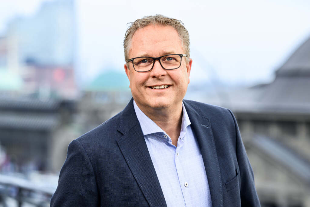 Ein Mann mit Brille, dunklem Blazer und hellblauem Hemd steht lächelt im Freien und im Hintergrund sind verschwommene Gebäude zu sehen.