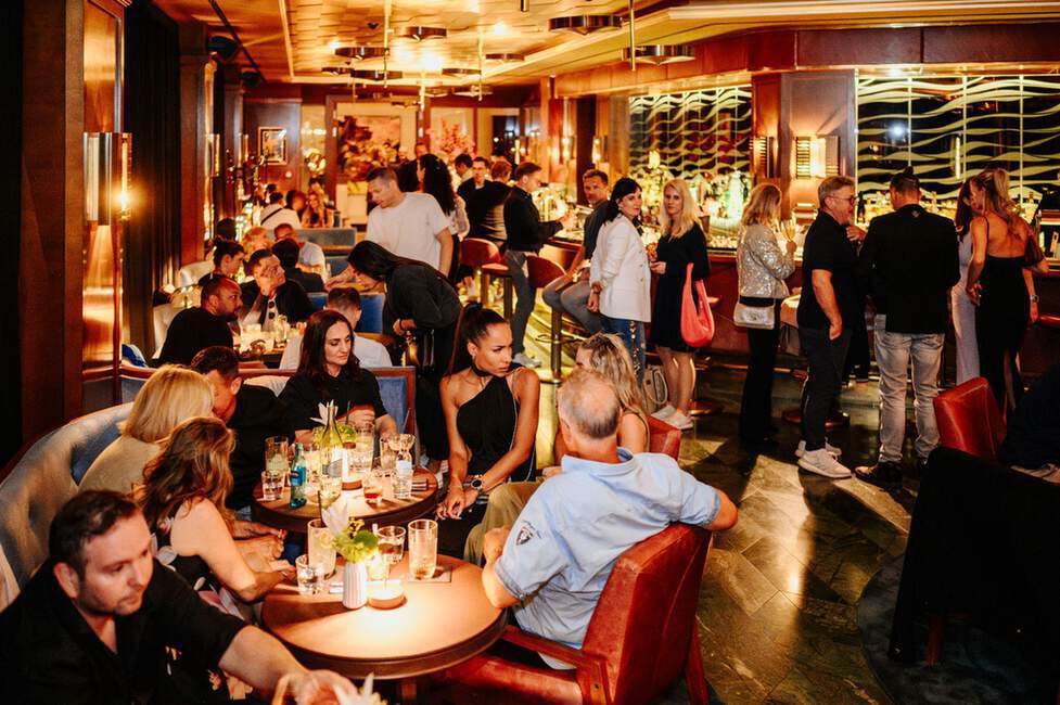 Eine gut besuchte Bar mit gedämpfter Beleuchtung und modernem Dekor, in der sich die Leute an den Tischen und im Stehen unterhalten.
