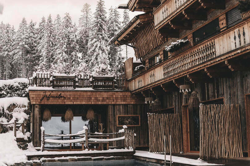 Ein verschneites Hotel nach traditioneller österreichischer Holzbauweise, mit Balkonen und Outdoor-Pool, umgeben von schneebedeckten Bäumen.