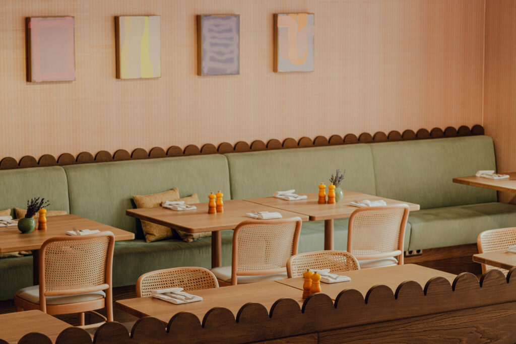 Das gemütliche Interieur der Alcron Bar Prague bietet Sitzgelegenheiten mit grünen Kissen sowie Tische und Stühle aus hellem Holz. Die Tische sind mit Servietten und Gewürzen gedeckt, während Kunstwerke die pfirsichfarbenen Wände schmücken und eine einladende Atmosphäre schaffen.