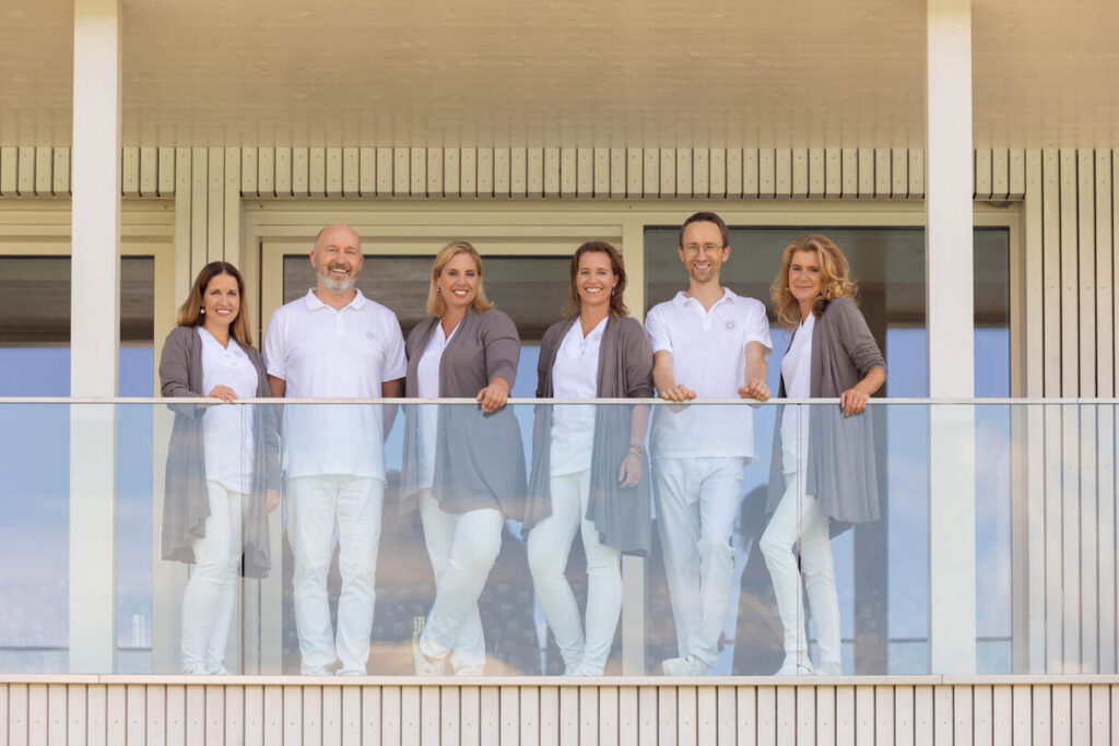 Sechs Menschen in weißer Kleidung und grauen Strickjacken stehen auf einem Balkon und lächeln in die Kamera. Dahinter ist ein Glasgeländer und helle vertikale Paneele.