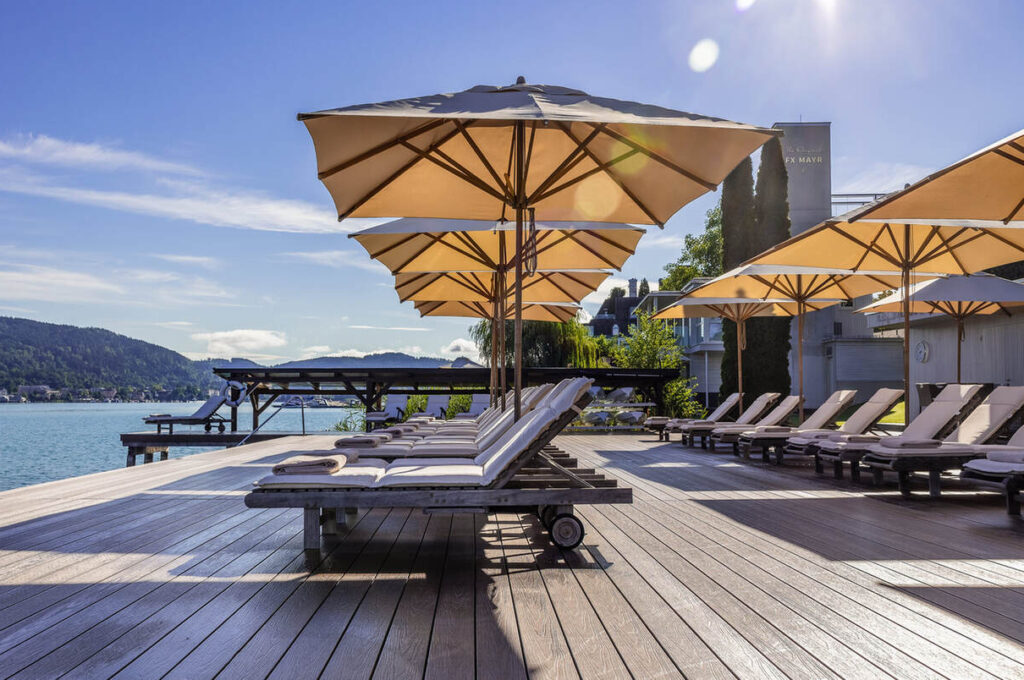 Sonnenliegen mit Sonnenschirmen auf einem Holzdeck mit Blick auf einen ruhigen See und einem klaren blauen Himmel im Hintergrund.