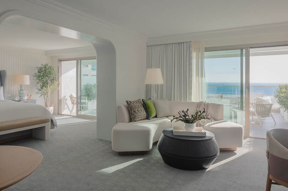 Modernes Hotelzimmer mit großem Bett, Sitzecke und Balkon mit Blick aufs Meer. Helle und neutrale Einrichtung mit rundem Couchtisch und raumhohen Fenstern.