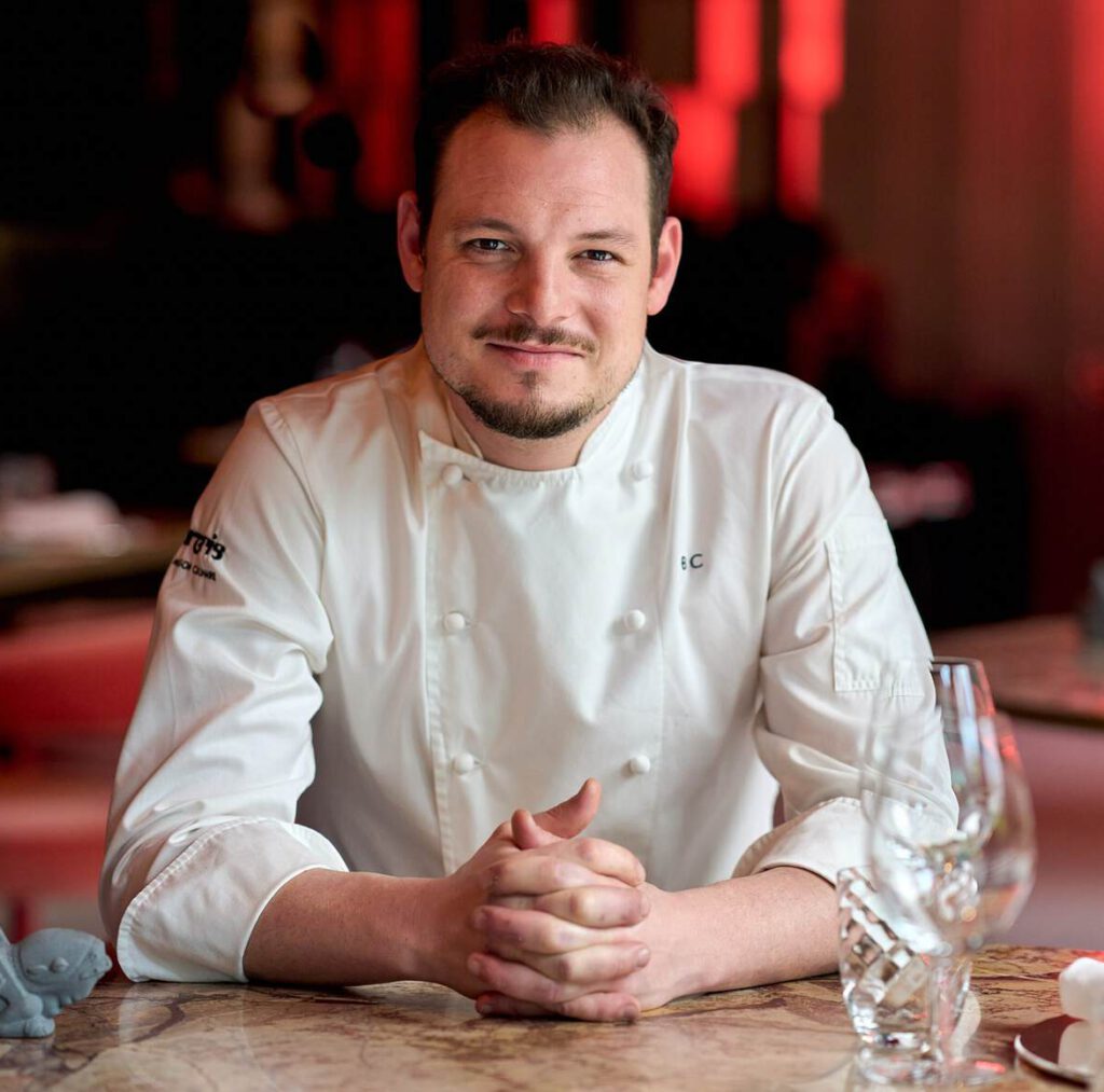 Ein Koch in weißer Uniform sitzt mit gefalteten Händen an einem Tisch, umgeben von Restaurantdekor und Glaswaren.