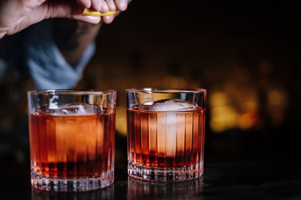 Zwei Gläser dunkelroter Cocktails mit Eiswürfeln stehen auf einer dunklen Fläche. Eine Hand presst eine Zitronenscheibe über eines der Getränke. Der Hintergrund ist unscharf.