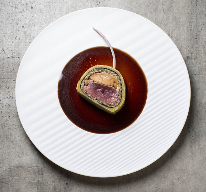 Ein Gericht auf einem weißen strukturierten Teller besteht aus einer in Teig eingewickelten Fleischscheibe, die mit Soße und einer dünnen Garnitur belegt ist. Der Teller steht vor einer grauen, strukturierten Oberfläche.